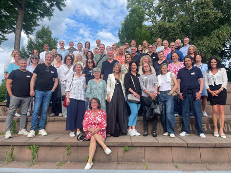 40 jahre Abitur Eine große Gruppe erwachsener Menschen steht und sitzt auf Steinstufen vor Bäumen und einem bewölkten Himmel.