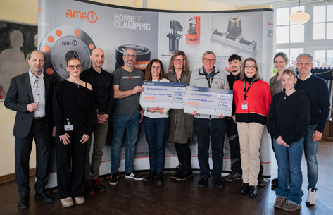 Azubi Projekt Eine Gruppe von etwa einem Dutzend Menschen steht vor einem Werbebanner und hält symbolische Schecks. Im Detail zeigt das Bild eine Gruppe von elf Personen, die sich vor einem Firmenbanner in einem Innenraum versammelt haben. Das Banner ist mit dem Logo