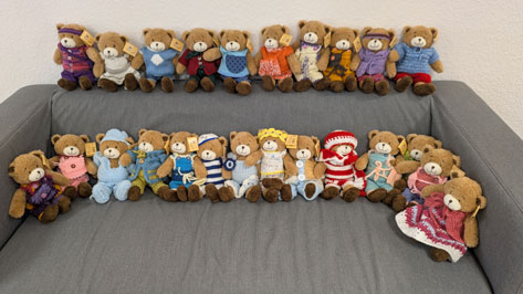 Bären Das Bild zeigt eine Sammlung von zwanzig Teddybären, die in zwei Reihen auf einem grauen Sofa sitzen. Jeder Bär ist mit einem einzigartigen gestrickten Outfit bekleidet, was der Ansammlung einen Hauch von Charme und Individualität verleiht. Die Outfits variieren in Farbe und Stil, von Pullovern und Overalls bis hin zu Mützen und Schals, was die Sammlung optisch ansprechend und gut zusammengestellt macht.