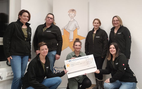 Das Bild zeigt sieben Frauen, die vor einem Roll-up-Banner mit dem Bild eines Kindes stehen. Sie halten einen Scheck der Volksbank Backnang eG über 500€ für den Verein „Dorfmädels ohne e.V.“ in den Händen. Die Frauen tragen einheitliche schwarze Jacken mit dem Logo „Dorfmädels“. Der Gesamteindruck ist der einer Spendenübergabe an eine gemeinnützige Organisation.