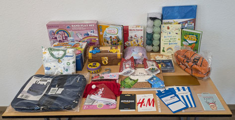 Das Bild zeigt eine große Auswahl an Geschenken und Spielzeug, die auf einem Tisch ausgebreitet sind. Es gibt Spielzeug für verschiedene Altersgruppen, darunter ein Sandkasten-Set, Bücher, einen Basketball, Kleidung und Dekorationsartikel. Die Artikel scheinen neu und verpackt zu sein, was darauf hindeutet, dass es sich um eine Sammlung von Geschenken handelt, vielleicht für ein Kind oder mehrere Kinder. Amazon und H&M Geschenkgutscheine sind ebenfalls zu sehen.