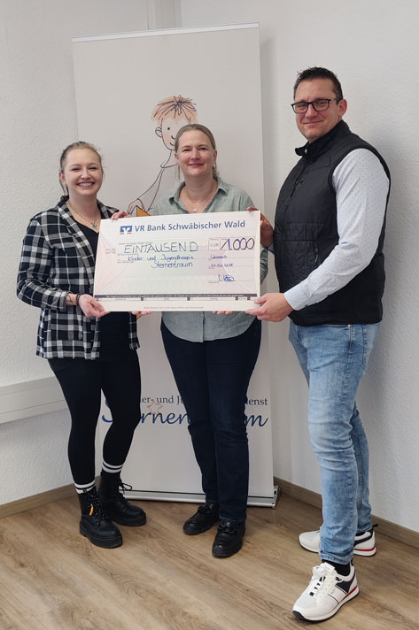 Das Bild zeigt drei Personen, zwei Frauen und einen Mann, die vor einem Roll-up-Banner der VR Bank Schwäbischer Wald stehen. Sie halten einen symbolischen Scheck über 1000 Euro in den Händen, der an das Kinder- und Jugendhaus Sternentraum geht. Die Szene wirkt positiv und dokumentiert eine Spendenübergabe. Das Bild strahlt Freude und den Erfolg einer gemeinschaftlichen Aktion aus.