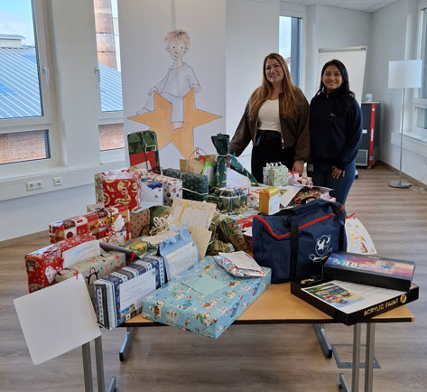 Helles Licht Das Bild zeigt zwei Frauen, die vor einem Tisch voller Geschenke stehen. Im Hintergrund ist ein Banner mit einer Zeichnung eines Kindes zu sehen, das auf einem Stern sitzt. Die Geschenke sind in verschiedenfarbiges Geschenkpapier eingepackt und scheinen für einen wohltätigen Zweck oder eine Weihnachtsaktion zu sein. Auf dem Tisch stehen außerdem eine blaue Tasche und ein Set Acrylfarben.