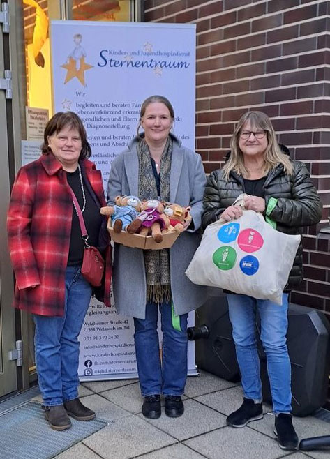 Das Bild zeigt drei Frauen vor einem Gebäude des Kinder- und Jugendhospizdienstes Sternentraum. Sie halten eine Kiste mit Stofftieren und eine Stofftasche. Die Frauen lächeln in die Kamera. Im Hintergrund ist ein Banner des Hospizdienstes mit dessen Kontaktdaten zu sehen. Das Bild vermittelt einen freundlichen und warmen Eindruck, vermutlich im Kontext einer Spendenübergabe oder einer ähnlichen Wohltätigkeit.