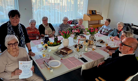 Eine Gruppe älterer Damen feiert mit zwei jüngeren Personen an einem gedeckten Tisch in einem hellen Raum.  Das Bild zeigt eine gesellige Runde von ungefähr zehn Personen. Die meisten Anwesenden sind ältere Damen, die um einen langen, weißen Tisch sitzen. Der Tisch ist festlich mit rosa Servietten, Sektgläsern, Blumenarrangements und verschiedenen Kuchen und Gebäcksorten gedeckt. In der Mitte des Tisches stehen Tulpen in Vasen. Eine ältere Dame sitzt vorne links und hält ein Dokument in den Händen, auf dem 