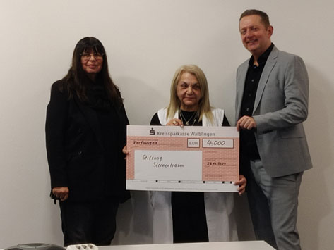 Das Bild zeigt drei Personen, die vor einem weißen Hintergrund stehen. Im Mittelpunkt steht eine Frau, die einen Scheck über 4.000 Euro von der Kreissparkasse Waiblingen für die Stiftung Sternentraum entgegennimmt. Links und rechts neben ihr stehen zwei weitere Personen, vermutlich Vertreter der Stiftung und der Sparkasse. Die Stimmung wirkt formell und positiv, es ist eine Übergabe-Szene mit dem sichtbaren Ergebnis einer Spende.