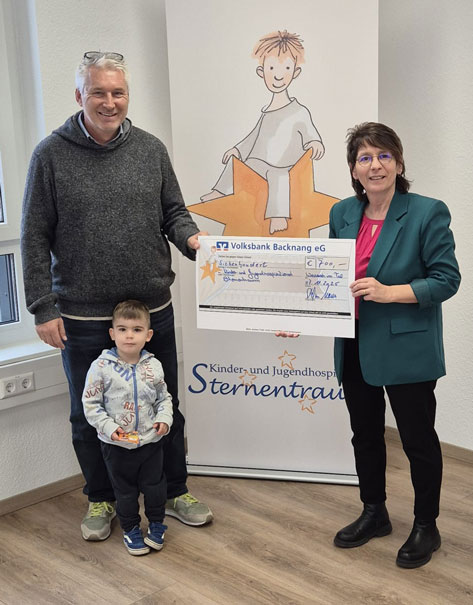 Moewes Das Bild zeigt drei Personen, die vor einem Roll-up-Banner für das Kinder- und Jugendhospiz Sternentraum stehen. Eine Frau in einem Blazer hält einen übergroßen Scheck der Volksbank Backnang eG über 700 Euro. Neben ihr stehen ein Mann und ein kleiner Junge. Der Mann trägt einen grauen Pullover, der Junge eine bedruckte Jacke und hält etwas Kleines in der Hand. Das Banner im Hintergrund zeigt eine Cartoon-Zeichnung eines Kindes, das auf einem Stern sitzt.