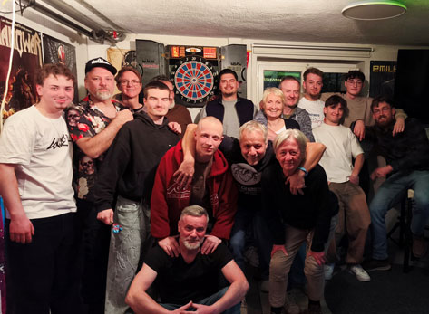 Dart Dieses Foto zeigt eine Gruppe von Personen unterschiedlichen Alters, die sich in einem gut beleuchteten Raum versammelt haben, wobei im Hintergrund eine Dartscheibe zu sehen ist. Die Szene ist im Querformat aufgenommen und die Gruppe steht eng zusammen, was ein Gefühl der Gemeinschaft und Vertrautheit vermittelt. Die Komposition ist leicht asymmetrisch, mit einer dichteren Ansammlung von Personen auf der linken und mittleren Seite des Bildes. Im Vordergrund kniet eine Person, während die anderen hauptsächlich stehen und sich teils umarmen oder die Arme um Schultern legen. Die Körpersprache ist insgesamt entspannt und positiv, was auf eine lockere Zusammenkunft hindeutet. Der Blickwinkel ist etwa auf Augenhöhe der meisten stehenden Personen, was eine direkte und persönliche Perspektive schafft. Die Personen auf dem Bild variieren in Alter und Geschlecht. Viele der Männer tragen Freizeitkleidung wie T-Shirts, Hoodies und Jeans. Ein Mann im Vordergrund, der kniet, hat einen kurz geschorenen Kopf und einen freundlichen Gesichtsausdruck, während er seine Hände vor sich gefaltet hat. Der Mann links daneben trägt ein schwarzes T-Shirt und scheint eine blonde Frau neben sich zu umarmen. Weitere Personen im mittleren Hintergrund zeigen unterschiedliche Gesichtsausdrücke, von Lächeln bis hin zu neutralen Blicken. Die Altersspanne reicht von jüngeren Erwachsenen bis hin zu älteren Personen. Kleidung und Frisuren sind vielfältig, aber sie alle scheinen Teil derselben informellen Gruppe zu sein. Das Foto ist eine digitale Fotografie. Der Stil ist realistisch und ungestellt, typisch für eine Momentaufnahme einer privaten Feier oder eines Treffens. Die technische Ausführung ist solide, mit guter Belichtung und Schärfe, die es ermöglicht, die Gesichter und Details der Personen gut zu erkennen. Es gibt keine offensichtlichen künstlerischen Effekte oder Filter, was dem Bild eine authentische und ungekünstelte Atmosphäre verleiht. Der Fokus liegt klar auf den Personen und ihrer Interaktion. Der Raum, in dem sich die Gruppe befindet, scheint ein privater Aufenthaltsraum oder eine Bar zu sein. An den Wänden hängen Poster, darunter eines, das wie ein Rock-Album-Cover aussieht. Eine Dartscheibe mit Pfeilen ist prominent im Hintergrund platziert, was auf Freizeitaktivitäten hindeutet. Die Beleuchtung ist hell und gleichmäßig, wahrscheinlich von Deckenleuchten, was die Szene gut ausleuchtet und keine dramatischen Schatten erzeugt. Die Umgebung wirkt gemütlich und einladend, was die positive Stimmung der Gruppe unterstreicht. Die Wände sind in einem hellen Farbton gehalten, und im Hintergrund sind auch Türen oder Fenster zu erkennen.