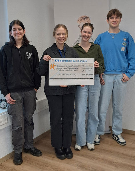 Das Bild zeigt vier junge Menschen, drei Frauen und einen Mann, die vor einer weißen Wand stehen. Eine der Frauen hält einen Scheck der Volksbank Backnang eG in der Hand. Der Scheck scheint eine Spende für einen guten Zweck zu sein, da darauf 