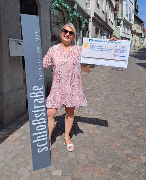 Sabine Kratter mit ihrem Geschäftsschild in der einen und einem großen Scheck in der anderen Hand auf der Straße