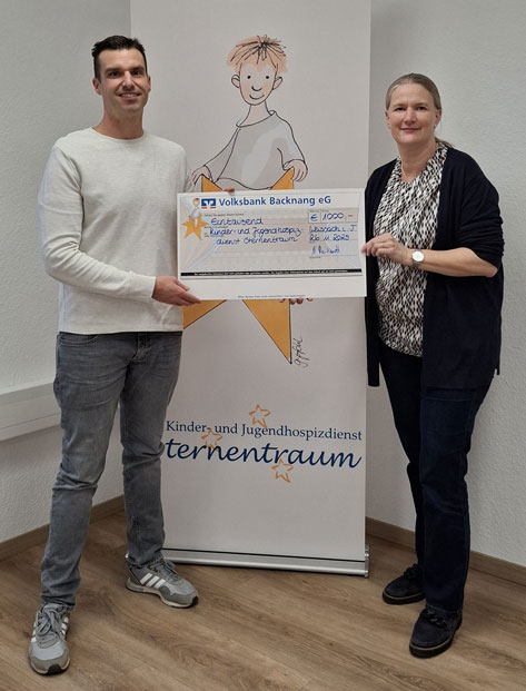 Das Bild zeigt zwei Personen, einen Mann und eine Frau, die nebeneinander stehen und einen überdimensionalen Scheck der Volksbank Backnang eG halten. Der Scheck ist über 1000 Euro ausgestellt und für den Kinder- und Jugendhospizdienst Sternentraum bestimmt. Im Hintergrund ist ein Roll-Up Banner mit dem Logo des Kinder- und Jugendhospizdienstes Sternentraum zu sehen, das ein kindliches Motiv und Sterne enthält. Beide Personen lächeln, was auf eine positive und freudige Stimmung hindeutet.