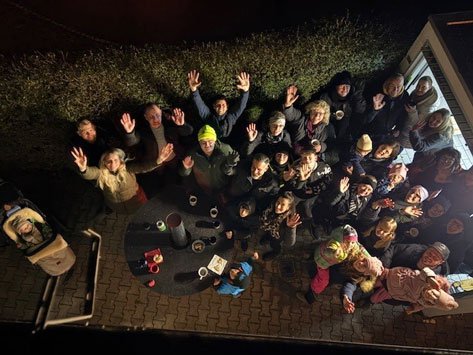 Spendenfest 2025_2 Das Bild zeigt eine Gruppe von etwa 30 Personen, die im Freien stehen und in die Kamera winken. Sie stehen auf einer gepflasterten Fläche und scheinen sich um einen Tisch zu versammeln, auf dem Tassen und andere Gegenstände stehen. Im linken Bildbereich befindet sich ein Kinderwagen mit einem Baby darin. Die Szene ist nachts aufgenommen und durch eine Lichtquelle erhellt, was eine warme Atmosphäre erzeugt.