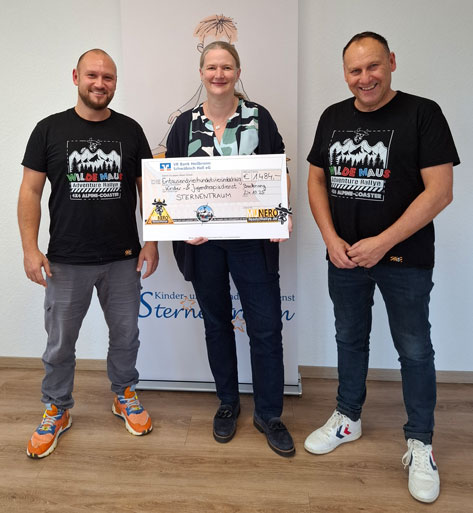 Wilde Maus Rallye Das Bild zeigt drei Personen, die anscheinend eine Spende überreichen. In der Mitte hält eine Frau einen übergroßen Scheck über 1.484 Euro. Auf dem Scheck steht der Name der Spendenorganisation „Sternentraum“. Die Personen links und rechts von ihr sind männlich. Alle drei lächeln in die Kamera. Sie stehen vor einem Banner, auf dem das Logo der Organisation „Kinder- und Jugenddienst Sternentraum“ abgebildet ist. Die beiden Männer tragen T-Shirts mit der Aufschrift „Wilde Maus Adventure Rallye Alpine-Coaster“.