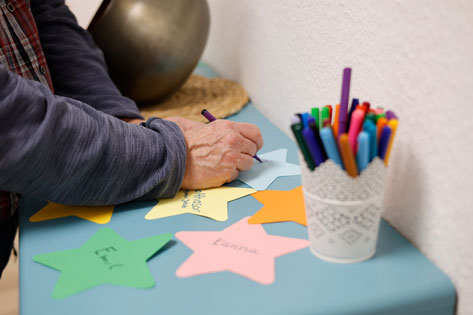 Bild Das Bild zeigt die Hände einer Person, die mit einem lila Stift auf einen gelben Stern aus Papier schreibt. Auf dem Tisch liegen weitere bunte Sterne, einige bereits beschriftet mit Namen wie